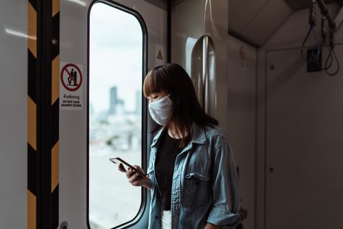 Free Asian woman wearing a face mask, using a smartphone on a train during the day. Stock Photo