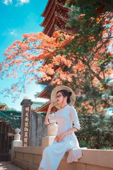 在日本寺庙附近，一名身穿白裙、头戴草帽的女子坐在樱花盛开的树下