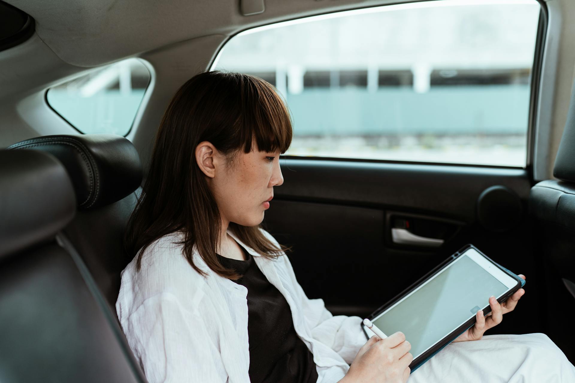 A focused Asian businesswoman works on a tablet from the backseat of a car. Urban lifestyle captured.