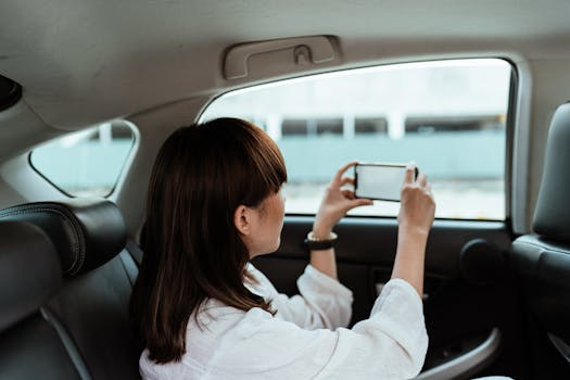 Side view of unrecognizable brunette in white shirt taking pictures on mobile phone with blank screen while riding in car backseat on daytime