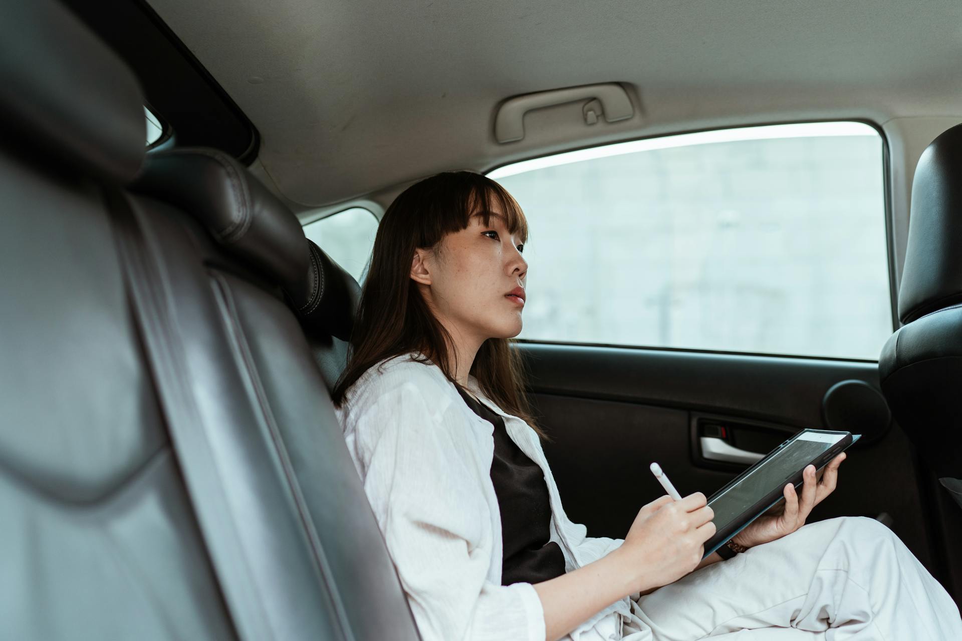 From below side view of contemplative Asian lady  in black and white outfit sitting in car passenger seat and working on tablet with stylus