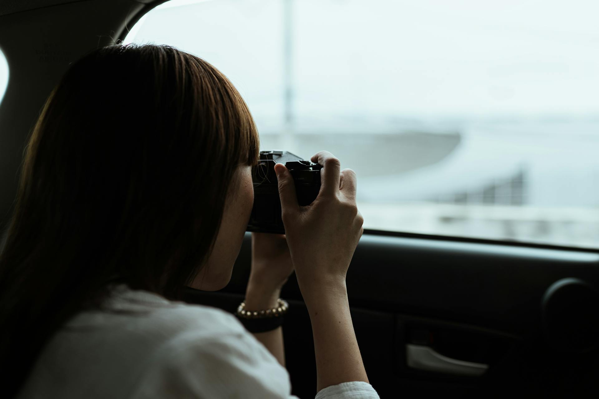 From behind anonymous brunette in white shirt taking pictures of urban buildings while riding in car passenger seat on daytime