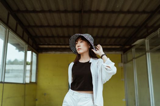 一位衣着时尚、头戴酷炫条纹帽的年轻亚洲女性，站在老建筑的黄色风化墙壁前，若有所思地移开目光，手轻轻触碰着帽子