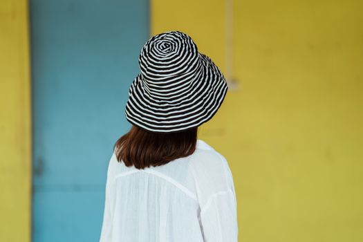 一名黑发女子头戴条纹帽，身穿白衬衫，背靠色彩缤纷的墙壁，显得活力十足又时尚
