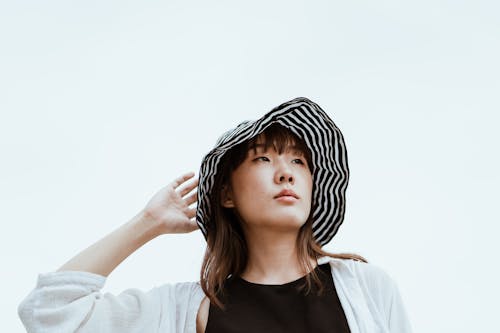 Low angle of thoughtful young Asian model in striped hat and casual wear looking away while standing leaned on hand near blue wall