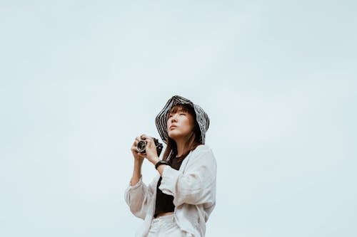 From below of young Asian female in striped hat and stylish wear looking away while standing with digital photo camera near blue wall