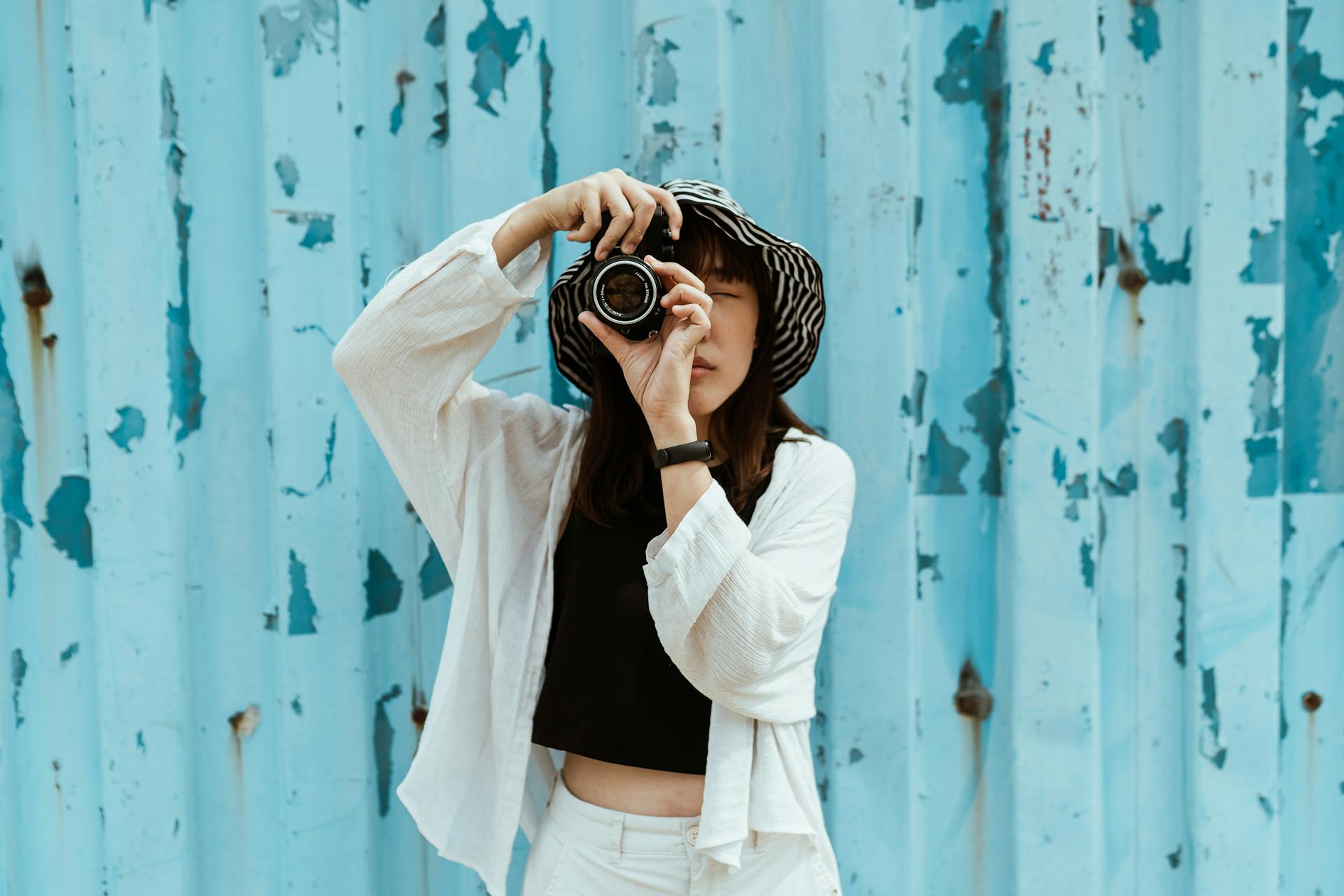 Asian female tourist in striped hat and casual clothes taking photo on professional photo camera while standing with closed eyes near blue ribbed wall with spots on surface