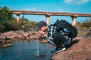 Side view of unrecognizable male traveler with rucksack drinking pure splashing water from river while squatting on rough shore