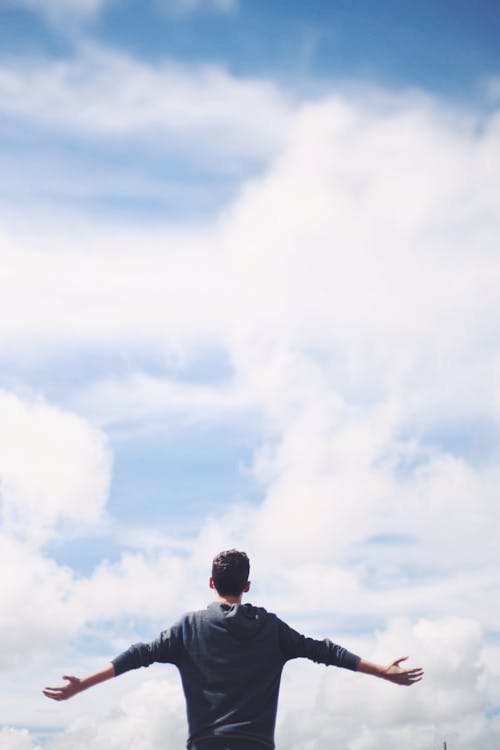 Free From below back view of anonymous male traveler standing with reached arms enjoying cloudy sky during vacation Stock Photo