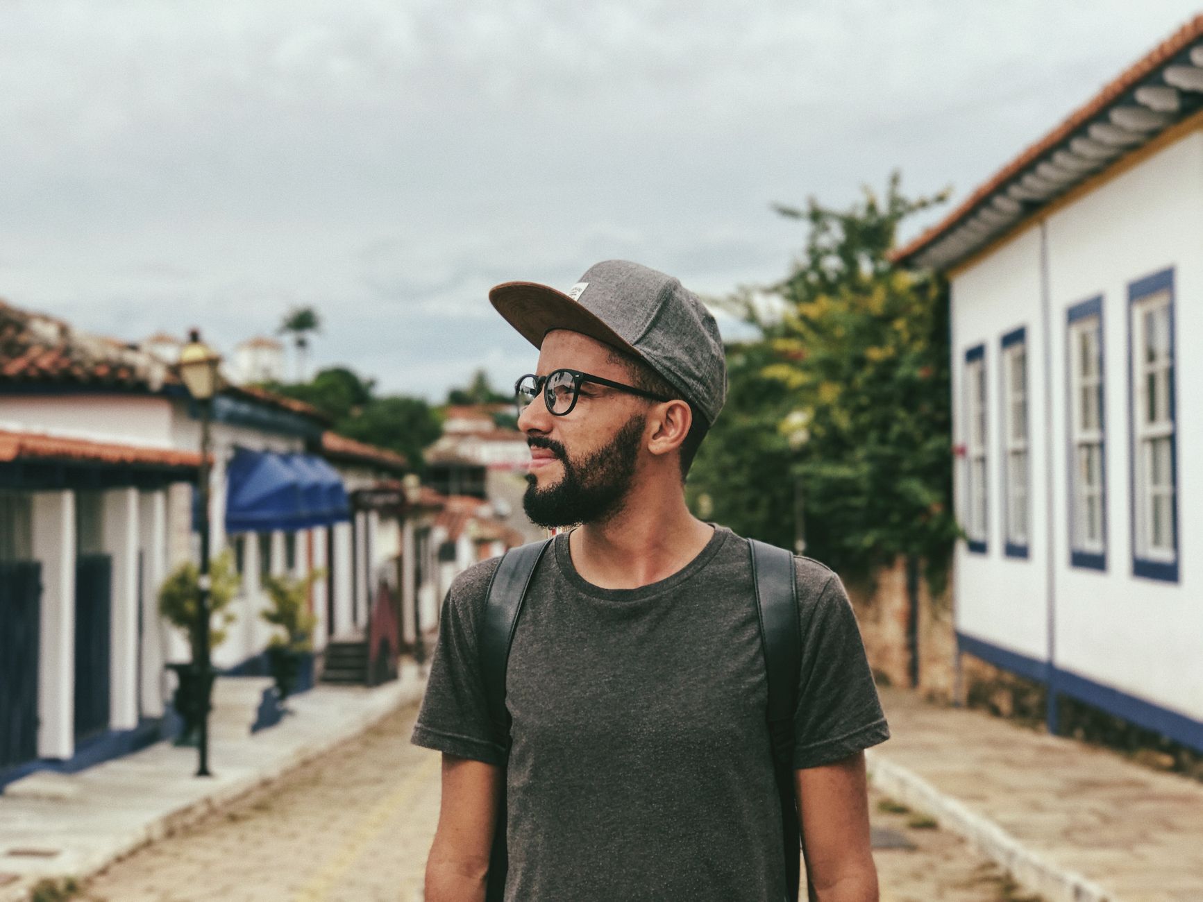 Young pondering ethnic male traveler in cap and eyewear looking away while standing on walkway near aged buildings