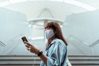 一名戴着口罩的亚洲妇女在现代室内楼梯上使用智能手机和耳机