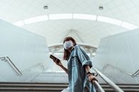 疫情期间，一名戴着口罩、身穿牛仔衬衫的妇女拿着智能手机和耳机站在楼梯上