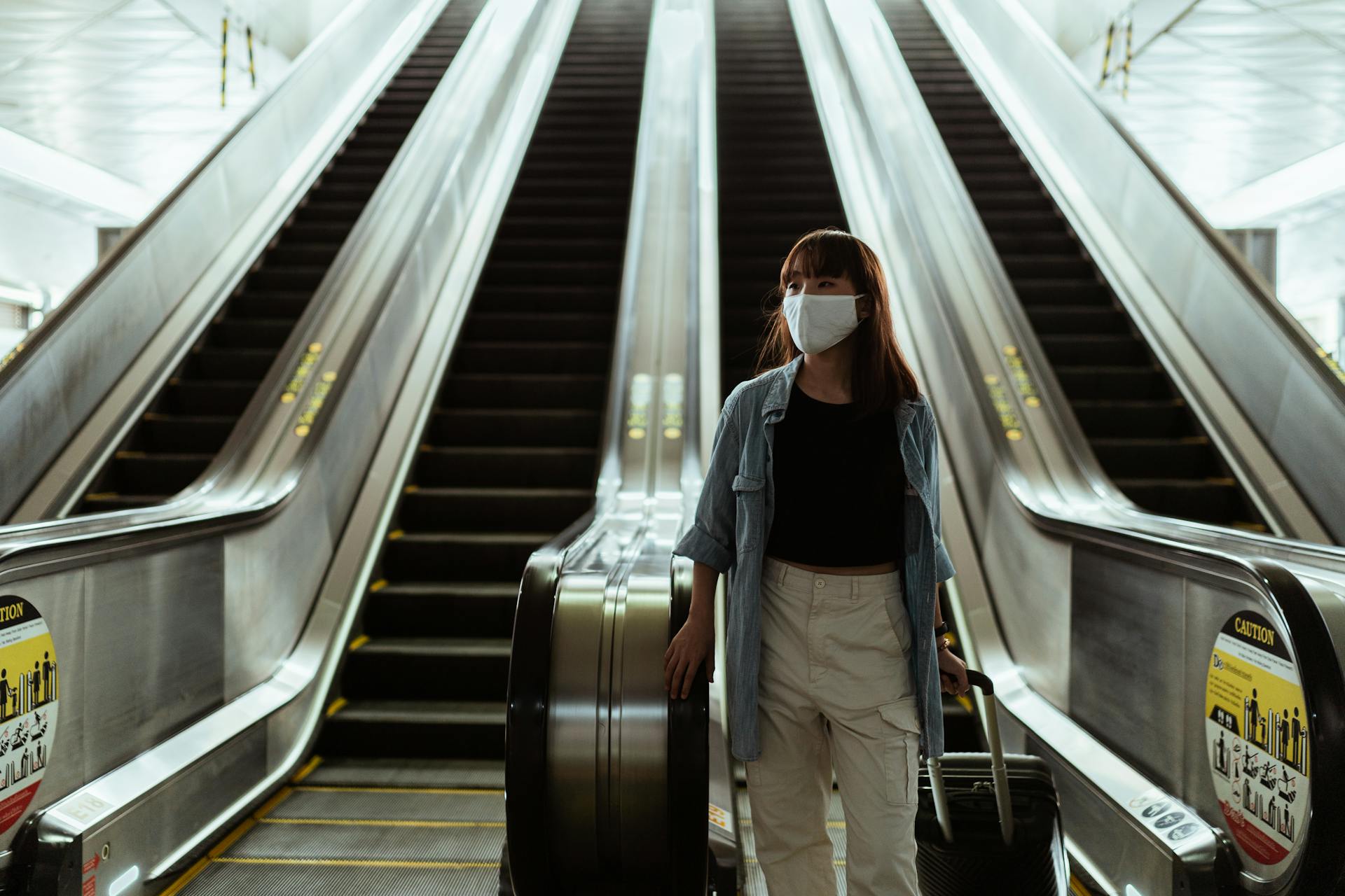 Asian woman wearing a face mask while traveling on an airport escalator during the pandemic, carrying luggage.