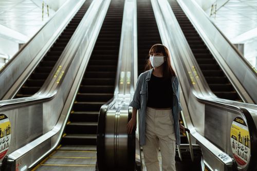 Free Asian woman wearing a face mask while traveling on an airport escalator during the pandemic, carrying luggage. Stock Photo