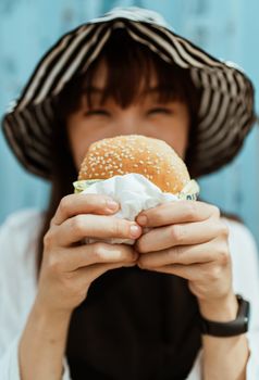 一位身穿休闲装、戴着智能手表的亚洲女性，面目全非地站在蓝墙附近，手里拿着美味的芝麻汉堡，正看着镜头