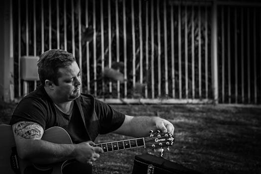 Monochrome image of a man tuning an acoustic guitar outdoors, showcasing musical focus.