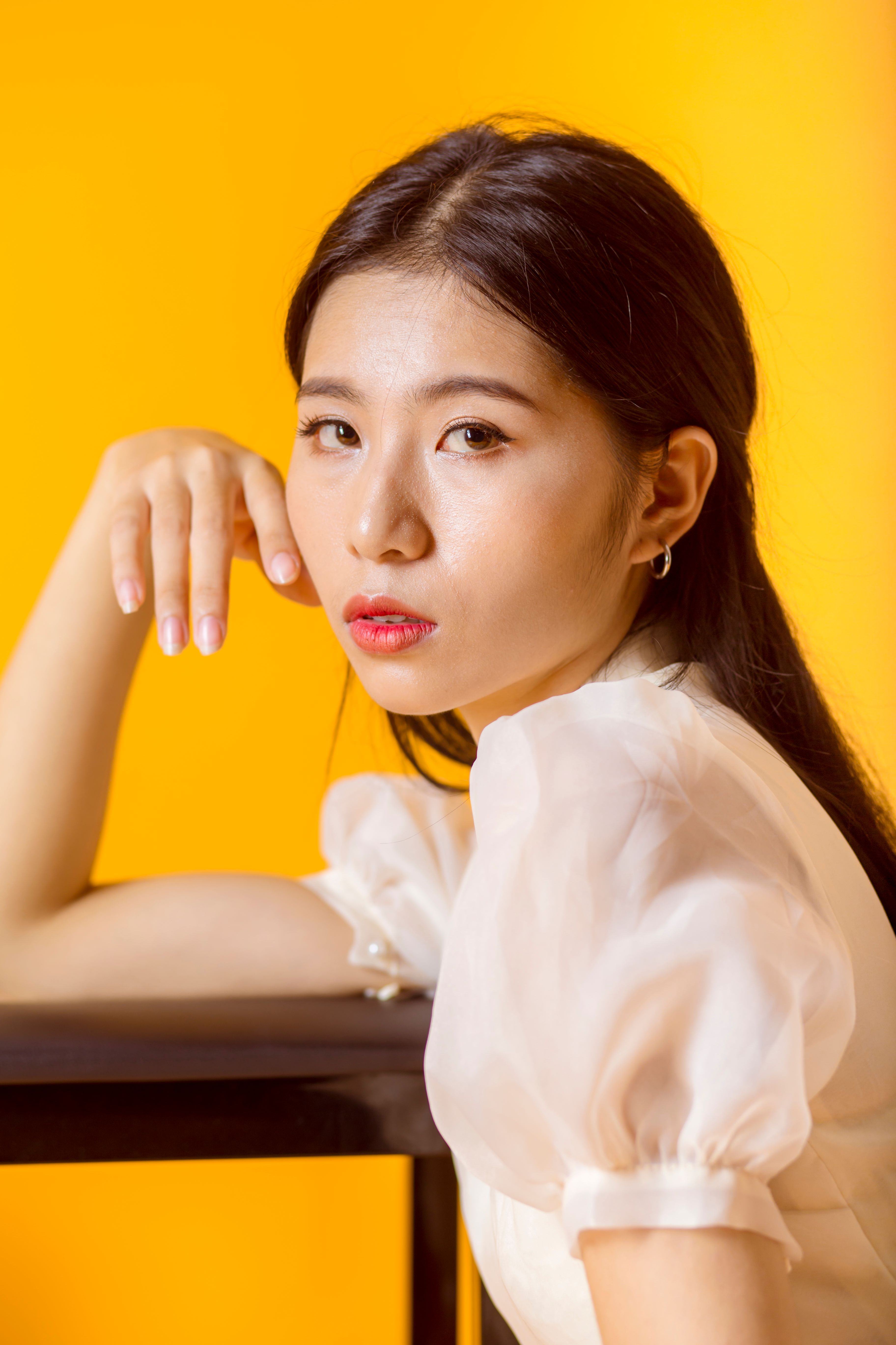 Beautiful Asian woman in white blouse posing with hand on face against a vibrant yellow background.
