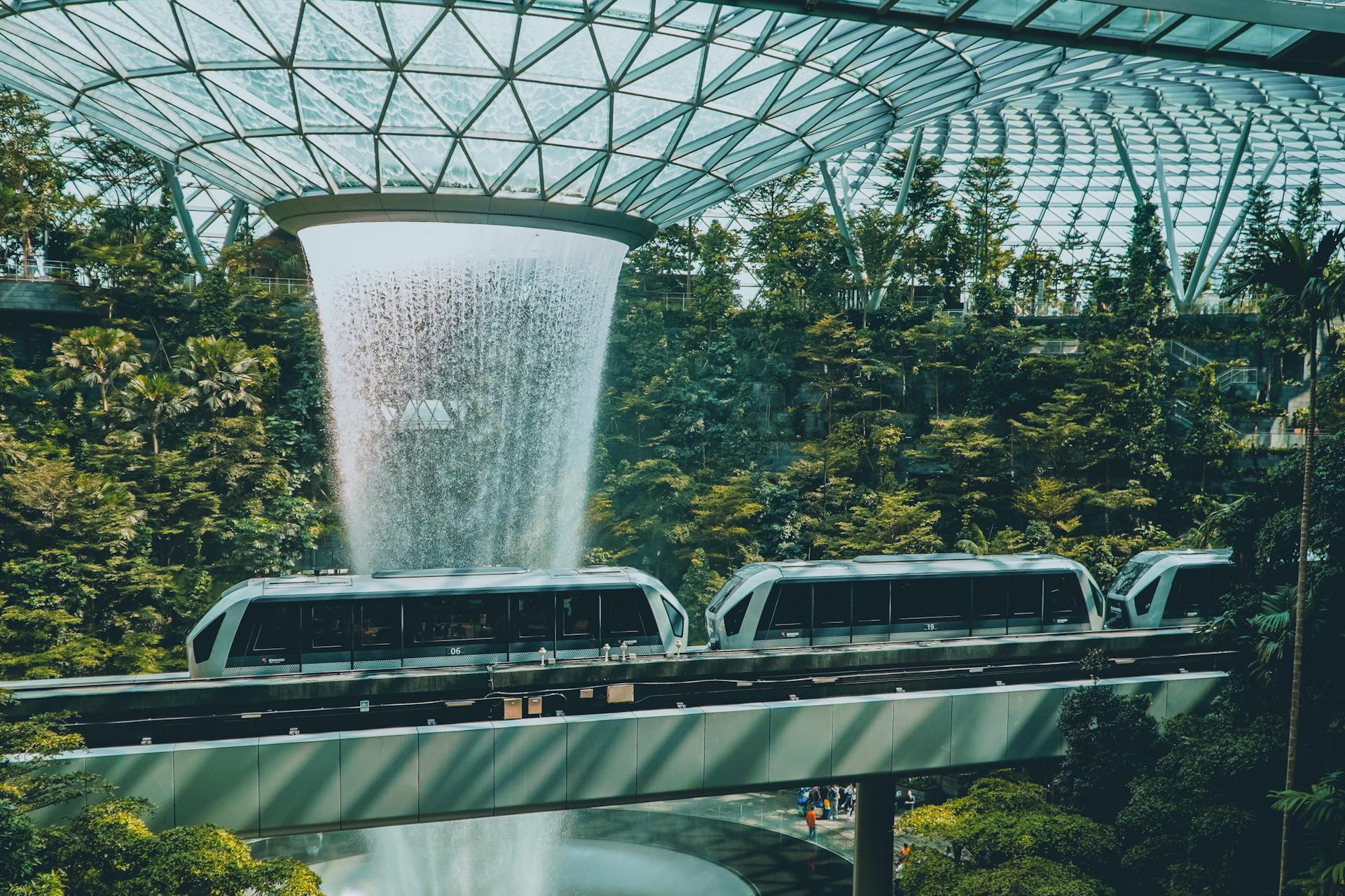 The stunning HSBC Rain Vortex waterfall at Jewel Changi Airport with a passing train and lush greenery.