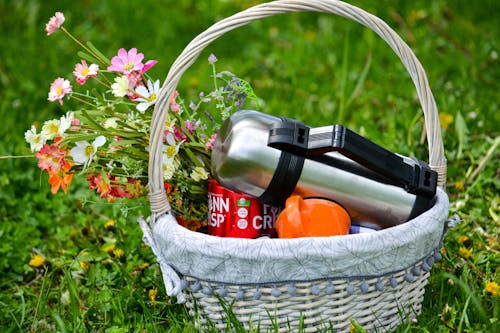 Wicker basket with cup and metal thermos near blooming flowers on bright green lawn in summer