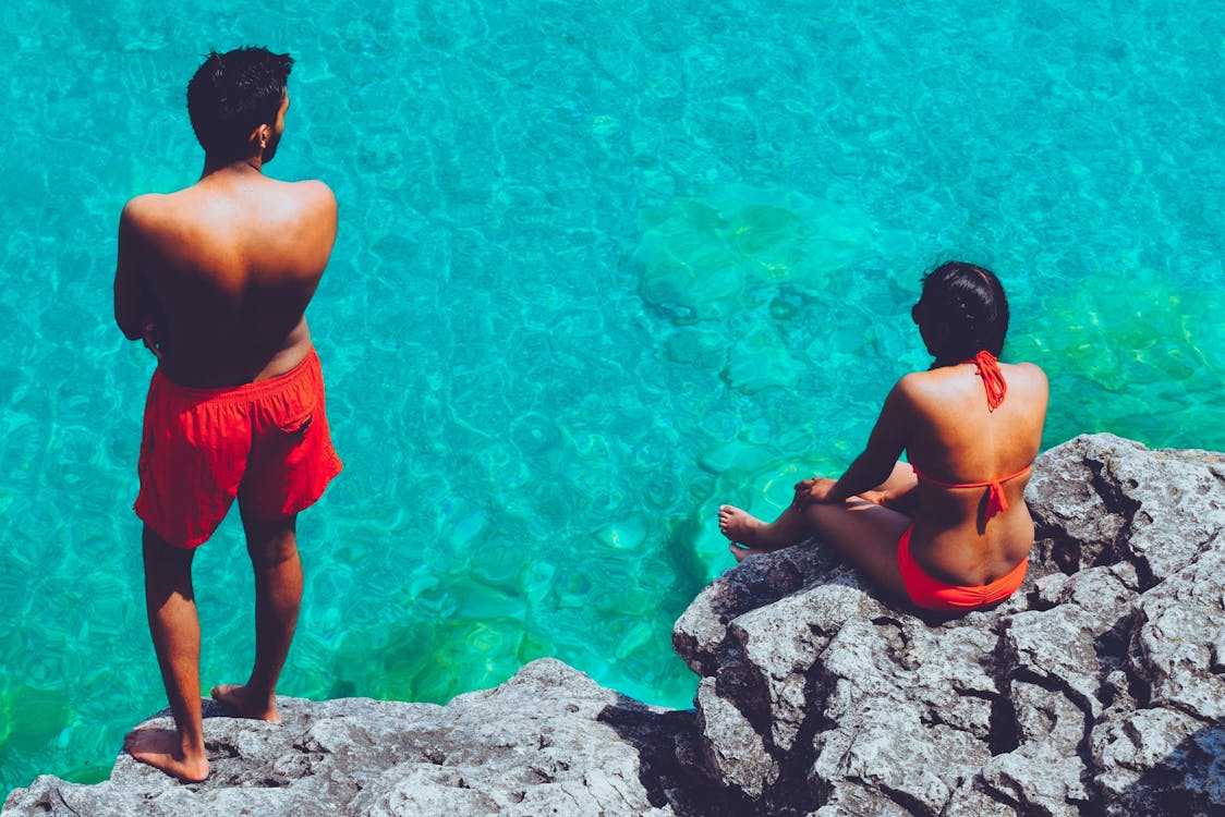 Free Man and Woman Standing and Sitting on Cliff Near Body of Water Stock Photo