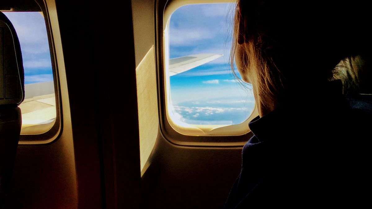 Silhouette of a woman looking out an airplane window at clouds and blue sky.