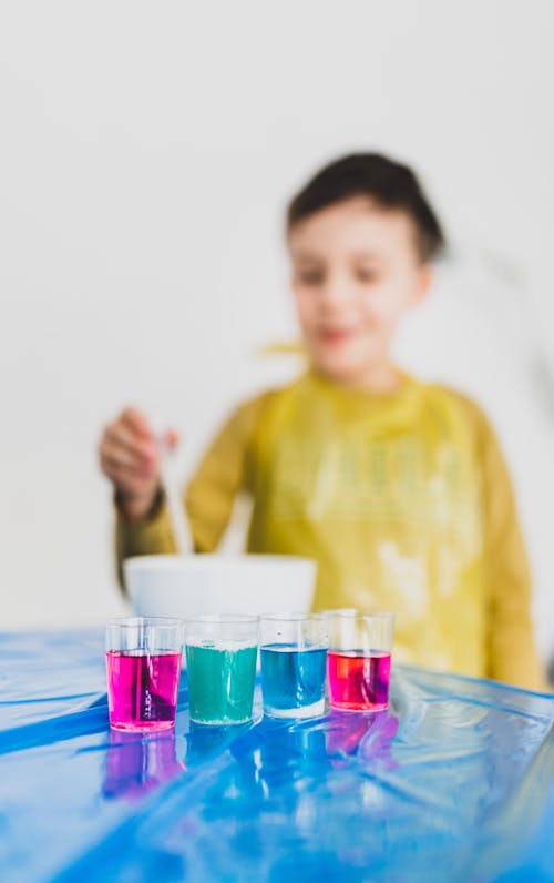 Cheerful little genius conducting chemical experiment near glasses with bright liquid substances in flat