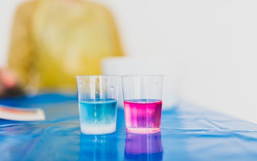 Faceless kid at table with transparent glasses with bright liquids on oilcloth during scientific experiment in house