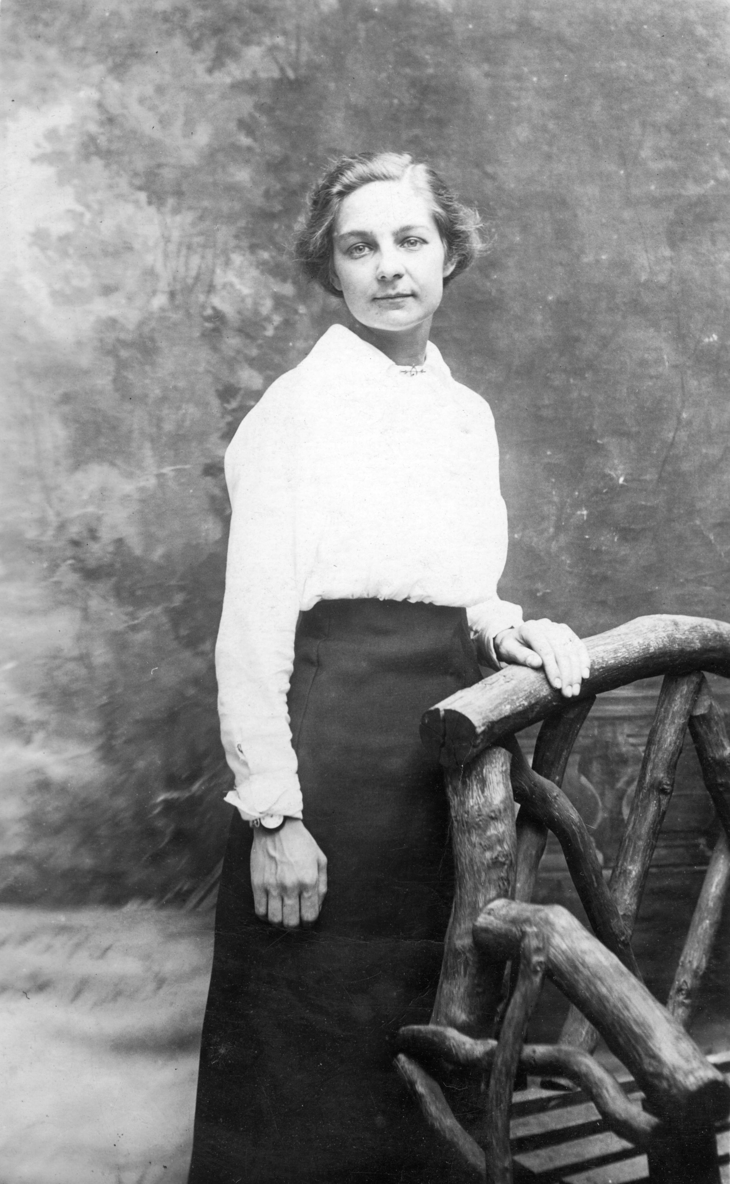 Elegant vintage portrait of a woman in classic attire standing by a rustic chair.