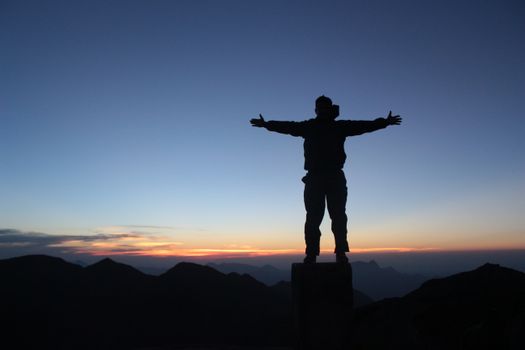 Back view of unrecognizable male traveler with outstretched arms enjoying sunset and nature on mountain peak