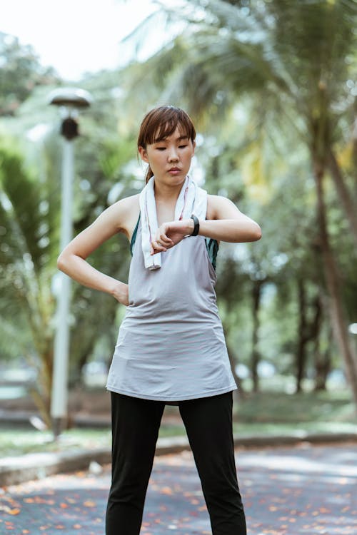 Slim Asian sportswoman in activewear and towel using wearable bracelet while standing with hand on waist on asphalt pavement in park in daylight