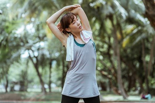 Fit young Asian female athlete with cotton towel in sportswear doing side bend while looking away in park with green trees in daylight