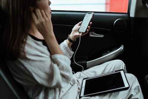 From above side view of crop anonymous female worker sitting in car with tablet and cellphone with blank screen while using earphones