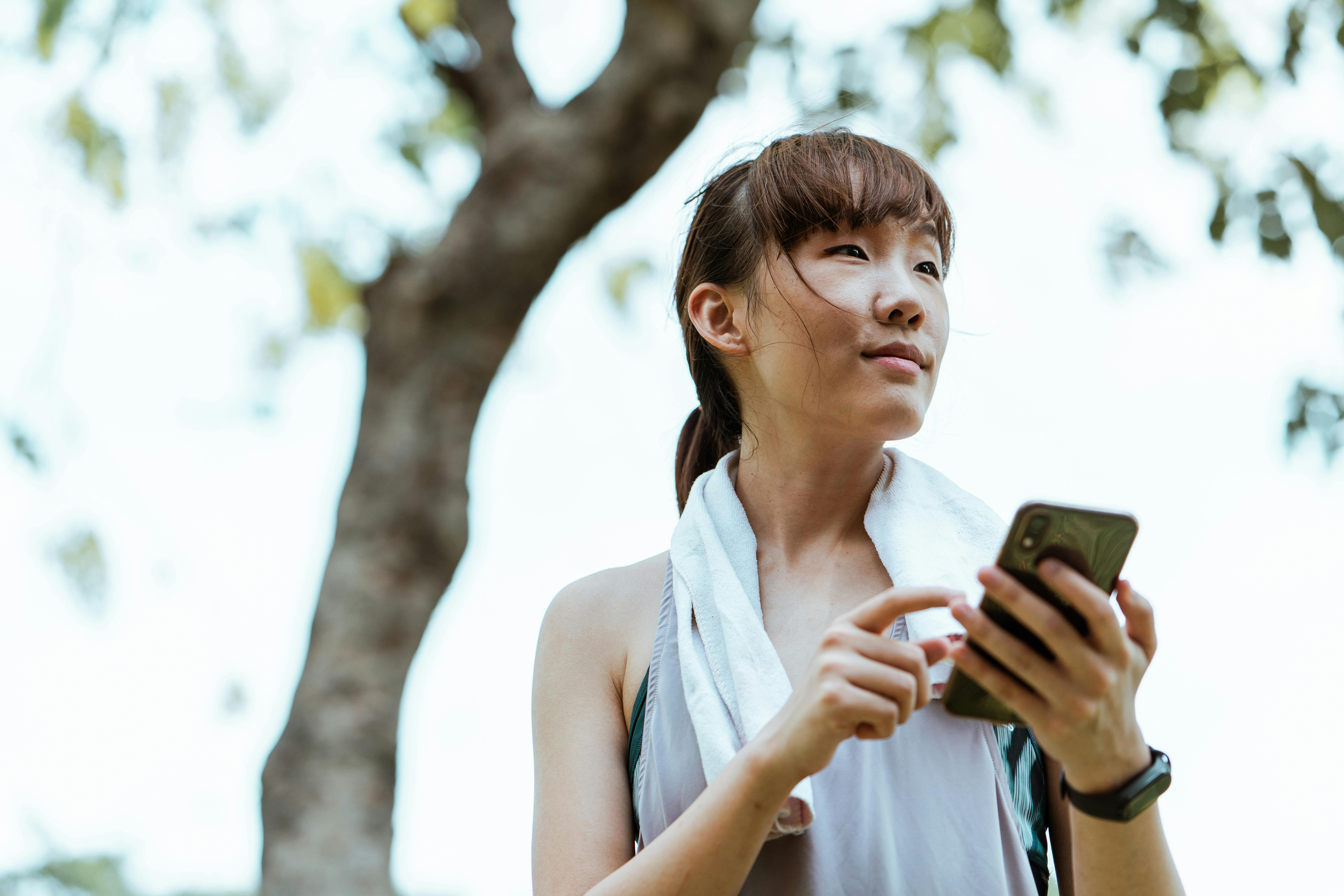 contemplative asian woman in bracelet browsing internet on smartphone in park