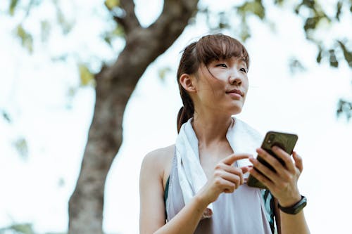 Contemplative Asian woman in bracelet browsing internet on smartphone in park