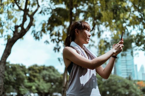 Free From below side view of slender Asian female athlete in sports clothes with towel taking selfie on mobile phone while standing near trees under sky and looking away Stock Photo