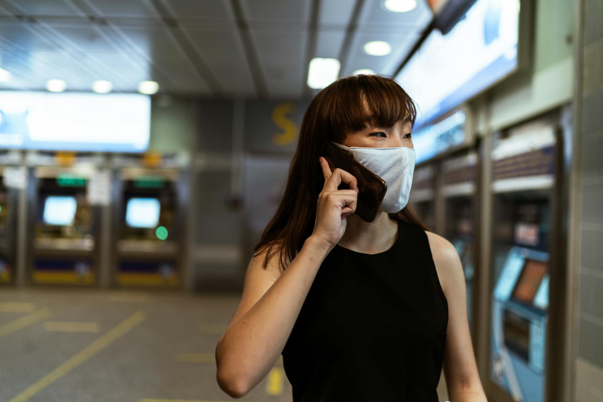 Asian woman talking on smartphone, wearing a face mask in a subway station, reflecting pandemic-era travel lifestyle.