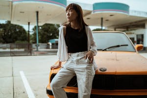 Slim pensive Asian woman in casual wear sitting with hand in pocket on old timer automobile parked on asphalt road and looking away