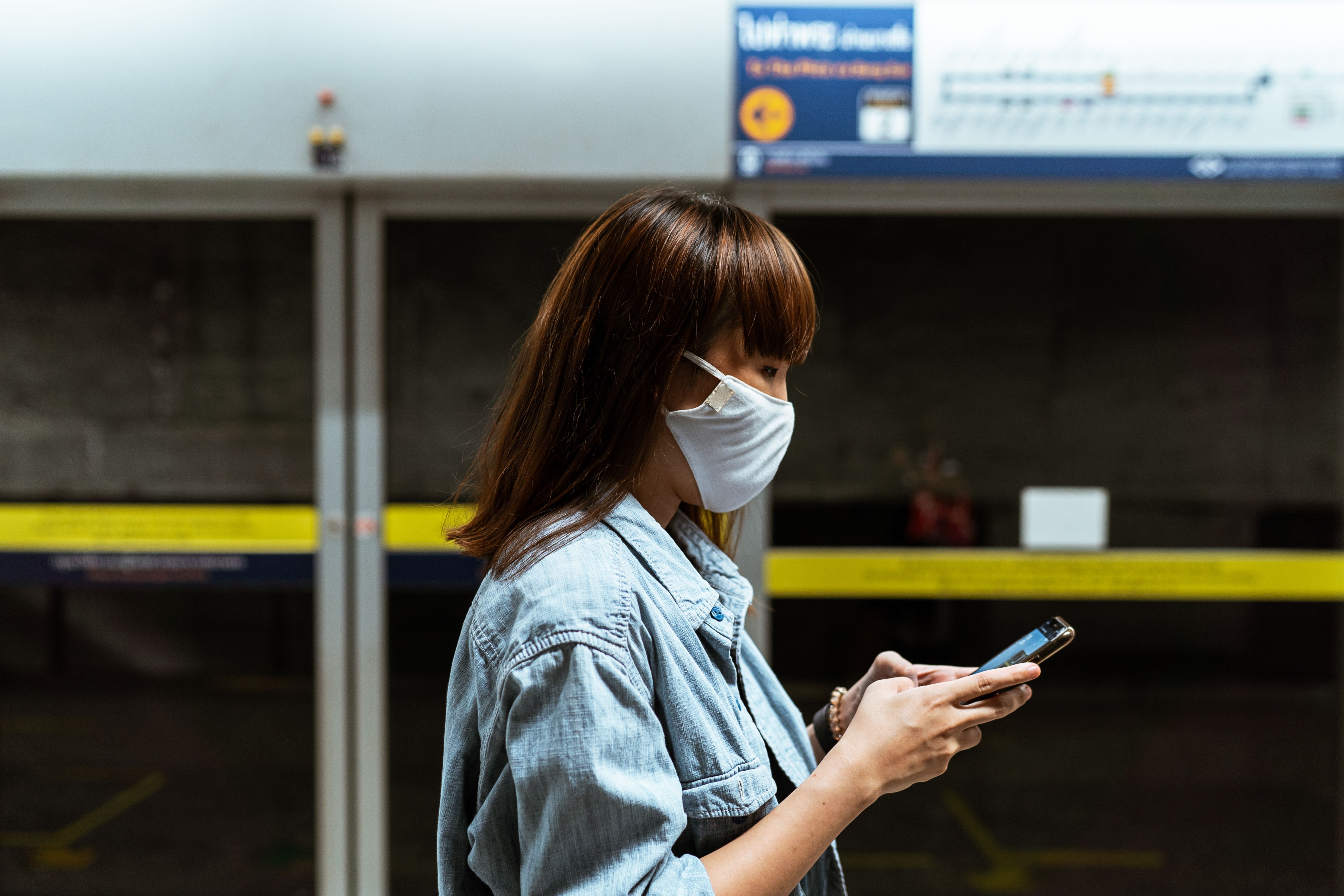 一名亚洲女子戴着口罩，在火车站室内用智能手机发短信