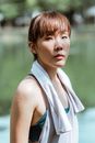 一位身材健美、怀揣梦想的亚洲女运动员，身着运动服，肩搭棉毛巾，在池塘边的公园休息，正望向镜头