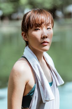 Side view of fit dreamy Asian female athlete in sports clothes with cotton towel on shoulders having break in park near pond and looking at camera