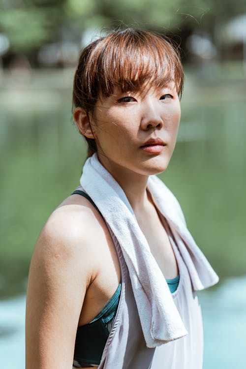 Free Side view of fit dreamy Asian female athlete in sports clothes with cotton towel on shoulders having break in park near pond and looking at camera Stock Photo