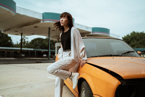 Slender young Asian female in casual wear looking away while standing leaned on one leg near old fashioned car parked on asphalt road on windy weather under cloudy sky