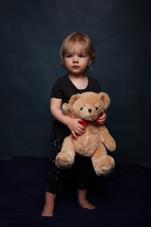 Free Adorable little girl in sleepwear standing on comfortable soft bed with teddy bear in hands Stock Photo