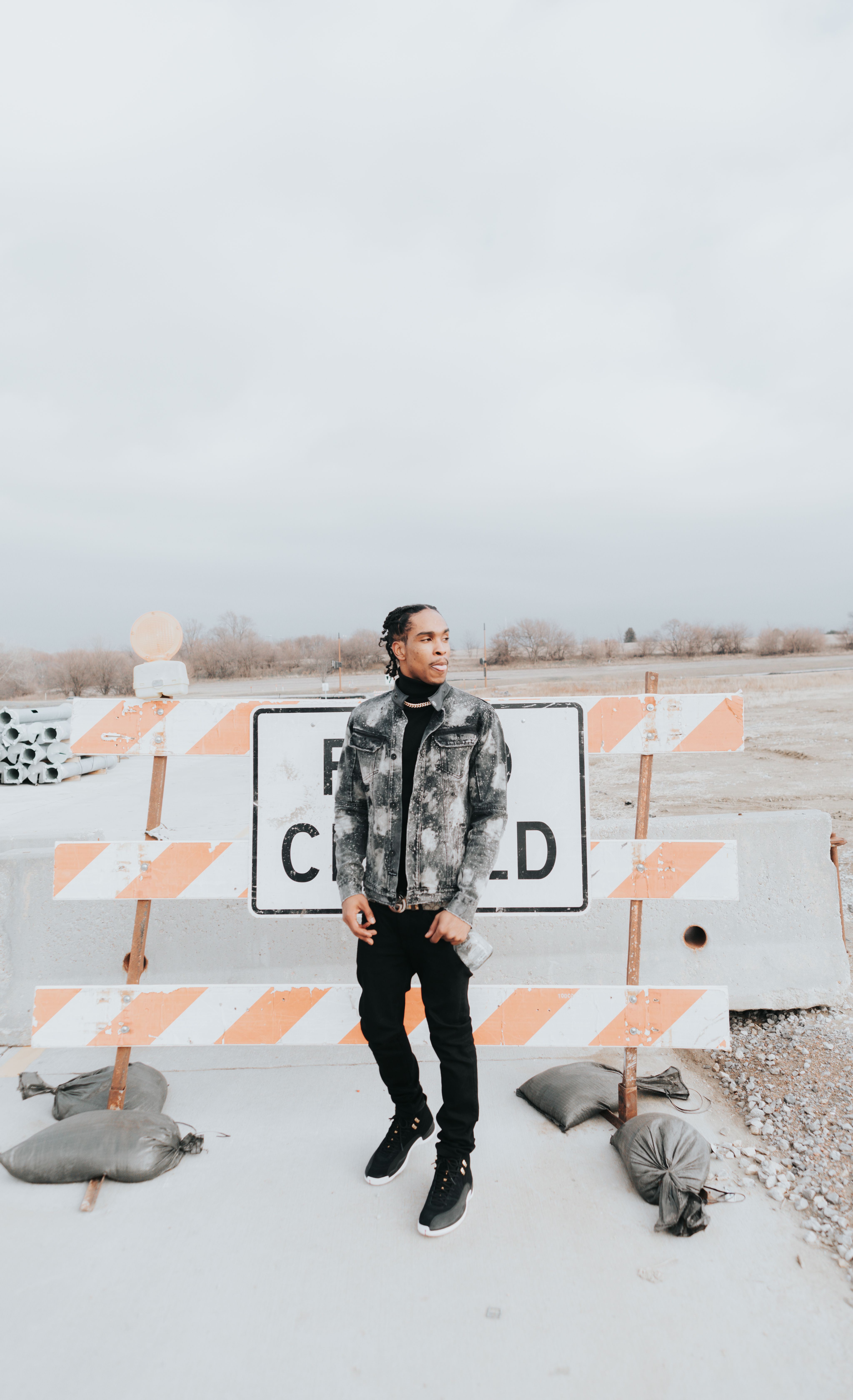 Man standing outdoors by a road closed sign, wearing casual attire and hands in pockets.