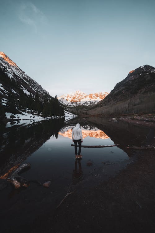 Back view of unrecognizable traveler in warm hoodie recreating on lake shore surrounded by snowy mountains against blue sky