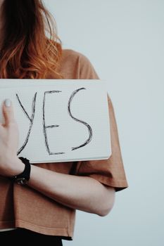 A woman wearing a watch holds a handwritten 'Yes' sign against a light background.