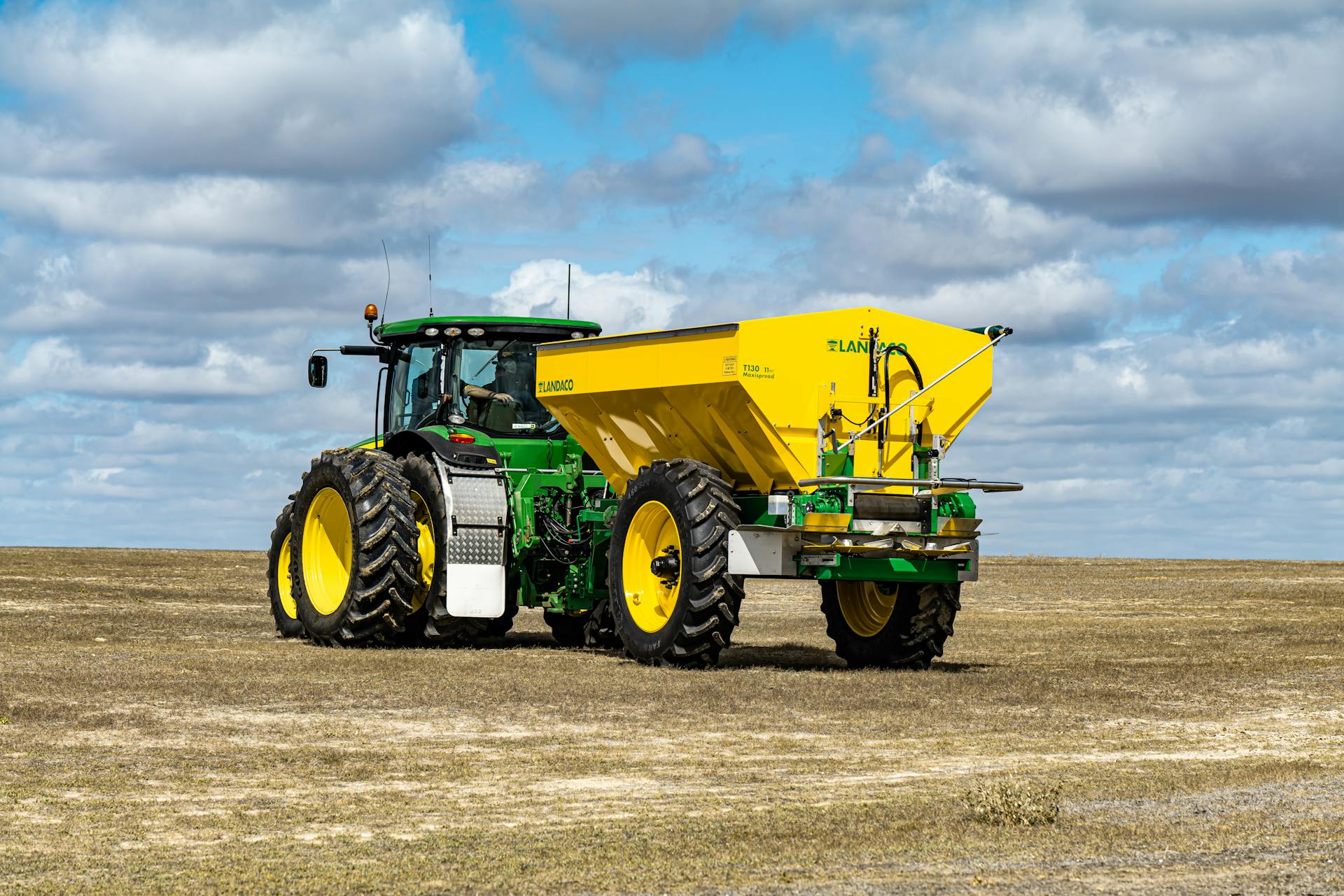 A green tractor with a yellow trailer on farmland under a bright blue sky with clouds.
