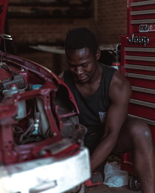 Positive young black male auto mechanic in comfortable clothes repairing vehicle during work in garage