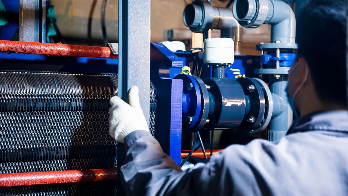 A worker in blue sleeves inspects pipes and equipment at an industrial plant