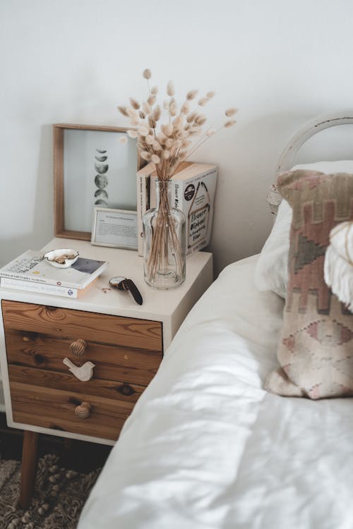 Free Cozy bedroom interior with wooden white bedside table with glass vase and books bear comfy bed with white sheets against white plain wall Stock Photo